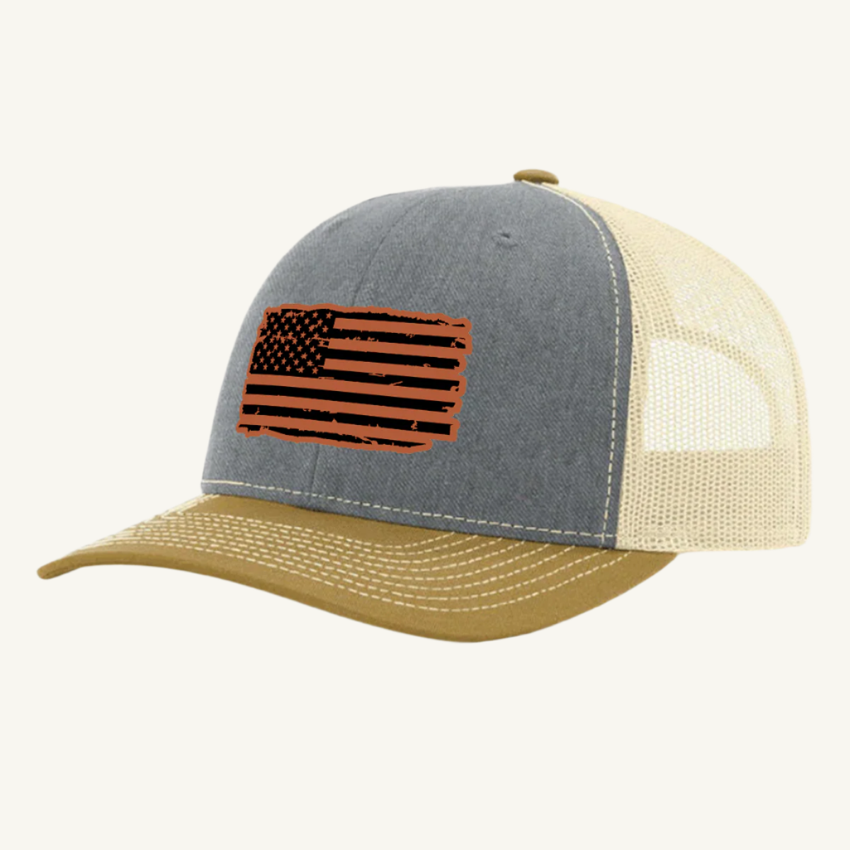 A heather gray, birch, and amber gold trucker hat with a distressed American flag design on the front, featuring a leatherette patch.