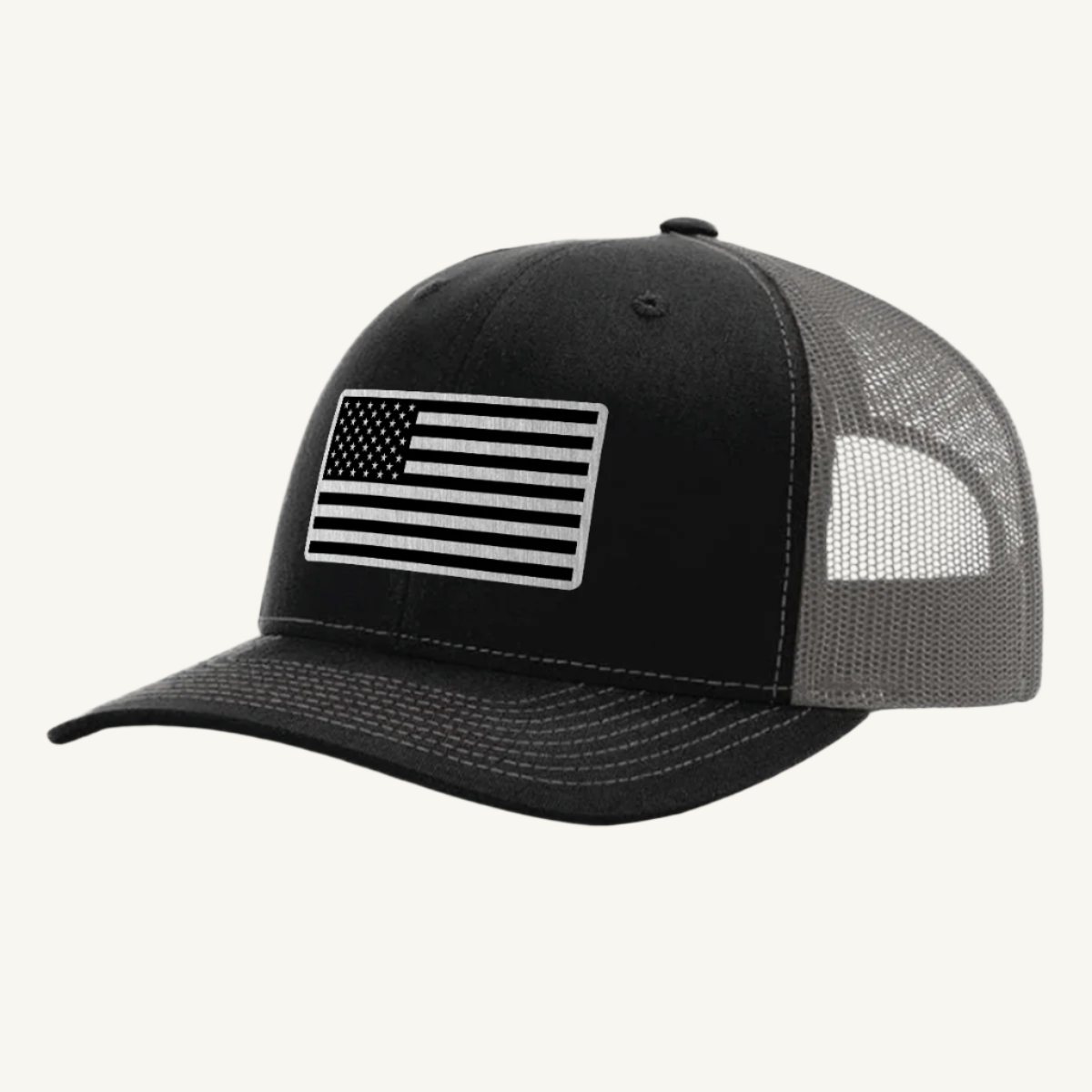 A black and charcoal trucker hat with a stainless steel acrylic patch featuring an American flag design on the front.