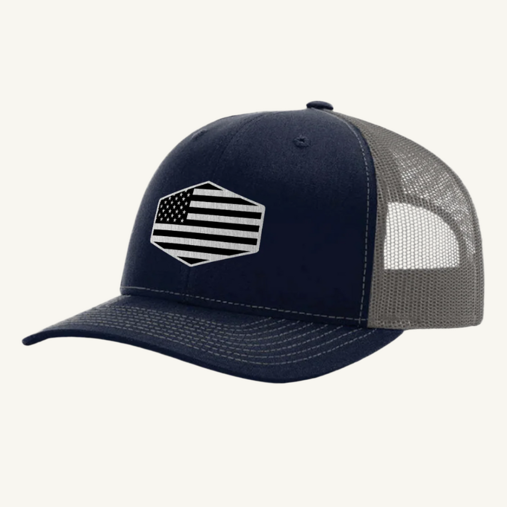 A navy blue and gray trucker hat with a stainless steel acrylic patch featuring an American flag design on the front.