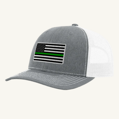 A heather gray trucker hat with an acrylic patch on the front displaying the Thin Green Line American Flag.