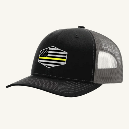 A black and charcoal trucker hat with a thin yellow line American flag acrylic patch on the front.